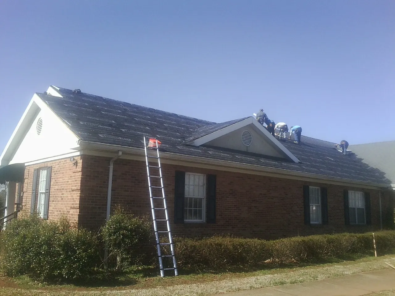 Crew tearing shingles off a brick ranch roof with a ladder set — Emergency Roof Repair work by our crew in Ray